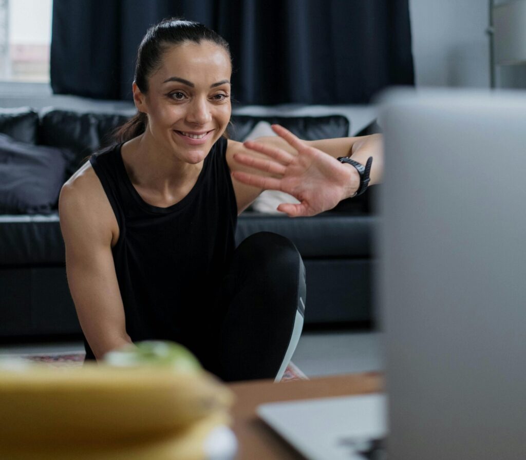 Woman in black fitness attire waving to a video call from home, smiling.